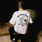 Person wearing a white t-shirt with 'The Dial Club' logo sitting on a stone wall.